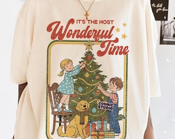 Könnte beinhalten: Cremefarbenes T-Shirt mit einer Retro-Illustration von Kindern, die einen Weihnachtsbaum mit einem Golden Retriever dekorieren. Der Text "It's the Most Wonderful Time" ist in Rot und Gold dargestellt. Das Shirt hat auch den Text "PNG SOLID & DISTRESSED".