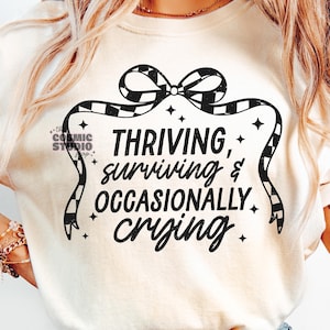 May include: White t-shirt with a black and white checkered bow and the text "THRIVING, surviving & OCCASIONALLY crying."