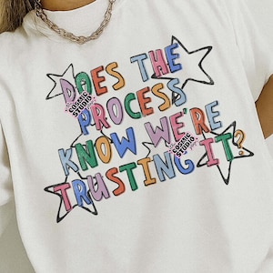 May include: White t-shirt with the text "DOES THE PROCESS KNOW WE'RE TRUSTING IT" in colorful, outlined letters and star shapes. The shirt also has the Cosmic Studio shop logo. The design is a digital download.