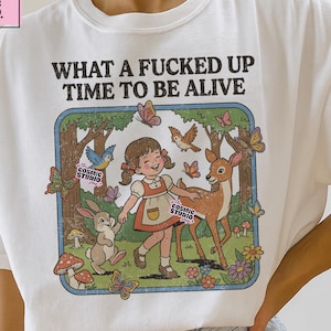 Puede incluir: Camiseta blanca con el texto "WHAT A FUCKED UP TIME TO BE ALIVE" sobre una colorida ilustración de una niña, un ciervo, un conejo, pájaros y mariposas. La camiseta también tiene el texto "the COSMIC STUDIO shop".