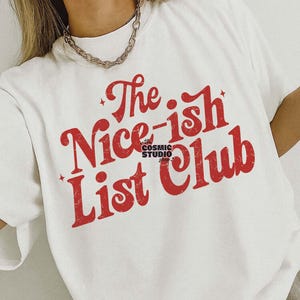 May include: White t-shirt featuring the red text "The Nice-ish List Club". The design includes stars and the Cosmic Studio logo. The shirt is a digital download and is available in three different colours.