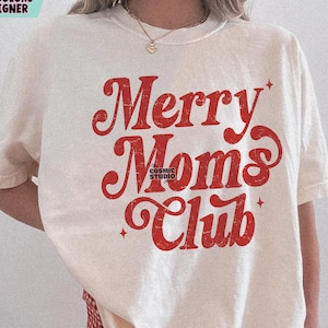 以下が含まれることがあります： クリーム色のTシャツ。赤色の文字で「Merry Moms Club」と書かれています。Tシャツはクルーネックで半袖です。下部にはCosmic Studio shopのロゴが見えます。