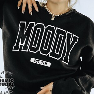 May include: Black sweatshirt with white text that reads "MOODY EST. 7AM". The Cosmic Studio shop logo is in the bottom left corner.