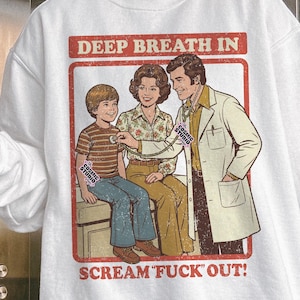 May include: White sweatshirt featuring a retro-style illustration of a doctor examining a child with a woman present. The text "DEEP BREATH IN" and "SCREAM FUCK OUT!" are displayed in a red frame. The Cosmic Studio logo is visible.