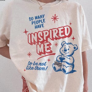 May include: Cream-colored t-shirt with the text "SO MANY PEOPLE HAVE INSPIRED ME to be not like them!" in red and blue. A blue teddy bear graphic holds a heart. The shirt has a distressed design.