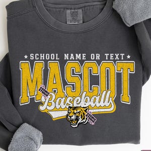 May include: A dark gray sweatshirt with the text "MASCOT Baseball" in yellow and white, with a tiger graphic. The text "SCHOOL NAME OR TEXT" is above the word "MASCOT". The sleeves are cuffed with a gray, fuzzy material.