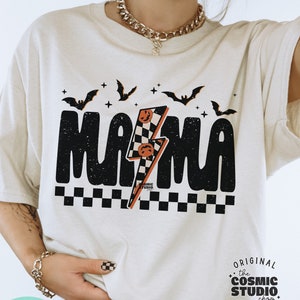 以下が含まれることがあります： 黒いグラフィックデザインが施された白いTシャツ。デザインには「MAMA」の文字と稲妻、市松模様が描かれている。グラフィックにはコウモリと星も含まれている。Tシャツの右下には円の中に「the COSMIC STUDIO shop. DESIGNER」の文字が書かれている。