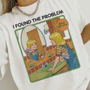 Puede incluir: Camiseta blanca con el texto "I FOUND THE PROBLEM" y una ilustración retro de una persona mirándose en un espejo. La ilustración presenta un baño verde y las palabras "Inner Reflection".