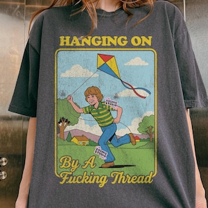 May include: Dark gray t-shirt featuring a vintage-style illustration of a boy flying a kite, with the text "HANGING ON By A Fucking Thread." The design includes a kite, boy, and landscape.
