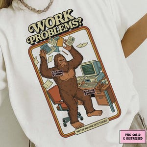 Może przedstawiać: Biała koszulka z grafiką w stylu retro przedstawiającą stworzenie podobne do Bigfoota przy biurku, otoczone papierami i komputerem. Tekst "WORK PROBLEMS?" znajduje się nad obrazem, a pod nim "NOT IF YOU'RE NOT THERE". Widoczne jest również logo Cosmic Studio.