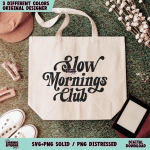 May include: A white tote bag with black text that reads "Slow Mornings Club". The text is in a retro font style. The bag is sitting on a green grassy surface.