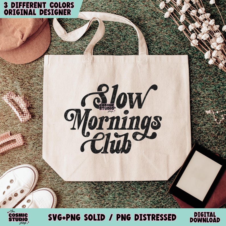 May include: A white tote bag with black text that reads "Slow Mornings Club". The text is in a retro font style. The bag is sitting on a green grassy surface.