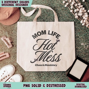 May include: A beige tote bag with the words "MOM LIFE Hot Mess Chaos Is Mandatory" in black script. The bag has cream-colored handles and is surrounded by other items, including a hat, shoes, and hair clips.