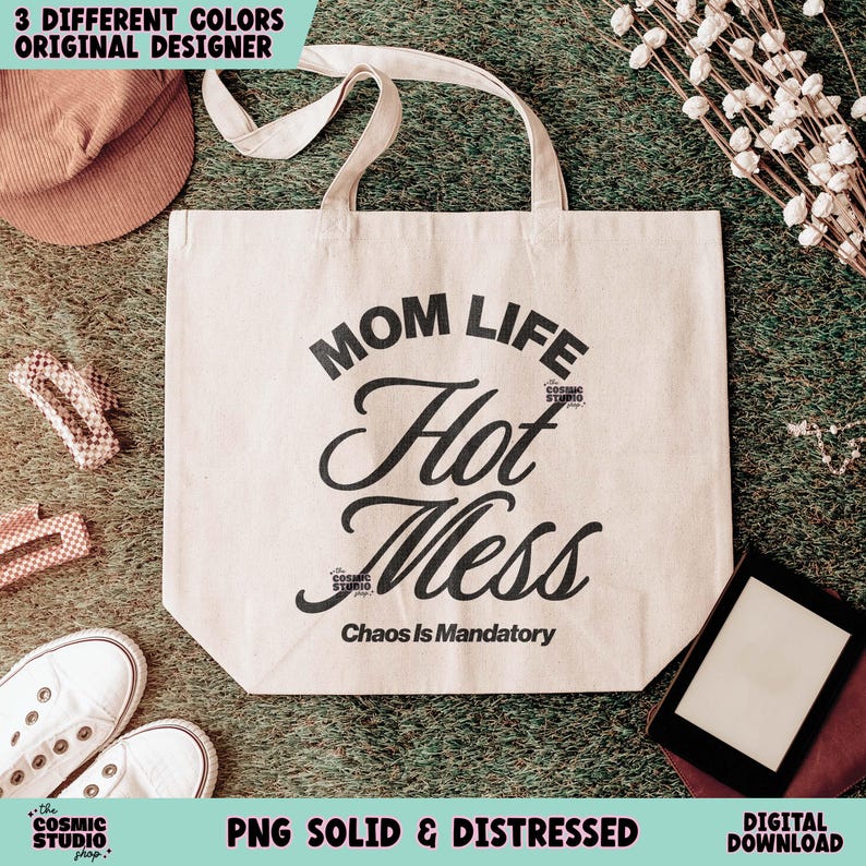 May include: A beige tote bag with the words "MOM LIFE Hot Mess Chaos Is Mandatory" in black script. The bag has cream-colored handles and is surrounded by other items, including a hat, shoes, and hair clips.