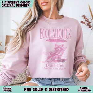 May include: Pink sweatshirt featuring a graphic of a raccoon reading a book under an umbrella on a beach. The text "BOOKAHOLICS" is above the graphic and "Beach Club Just one more chapter" is below the graphic.