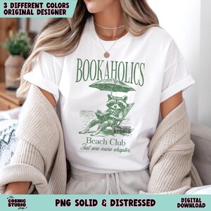 May include: White t-shirt featuring a green graphic of a raccoon reading a book under an umbrella. The text "BOOKAHOLICS" is above the raccoon and "Beach Club" and "Just one more chapter" are below the raccoon.