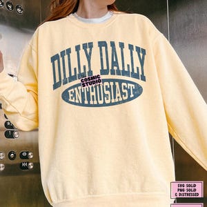 K&ouml;nnte beinhalten: Ein blassgelbes Sweatshirt mit Rundhalsausschnitt und dunkelblauem Schriftzug im College-Stil, der DILLY DALLY ENTHUSIAST lautet. Das Design weist eine abgenutzte Textur in den Buchstaben und eine horizontale ovale Form um das Wort ENTHUSIAST auf.