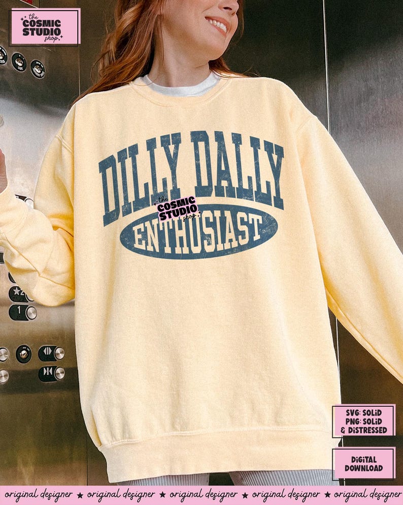 K&ouml;nnte beinhalten: Ein blassgelbes Sweatshirt mit Rundhalsausschnitt und dunkelblauem Schriftzug im College-Stil, der DILLY DALLY ENTHUSIAST lautet. Das Design weist eine abgenutzte Textur in den Buchstaben und eine horizontale ovale Form um das Wort ENTHUSIAST auf.