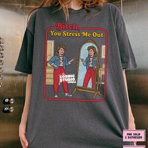 以下が含まれることがあります： 赤と黄色の「Bitch, You Stress Me Out」という文字がプリントされた濃いグレーのTシャツ。鏡に映った自分を指さす女性のグラフィック。Cosmic Studioのロゴが見えます。デジタルダウンロード可能。
