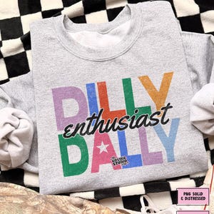 May include: A heather gray sweatshirt with the text "DILLY enthusiast DALLY" in colorful block letters. The design includes a star and the words "the COSMIC STUDIO shop". The sweatshirt is a digital download.