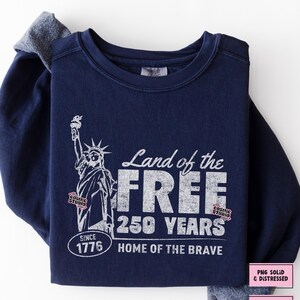 以下が含まれることがあります： 自由の女神と「Land of the Free, 250 Years, Home of the Brave, Since 1776」の文字がプリントされた、白のグラフィックが施された濃い青色のスウェットシャツ。クルーネックと長袖です。