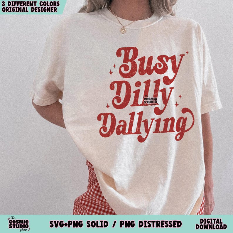 May include: Cream-colored t-shirt with the phrase "Busy Dilly Dallying" in distressed red lettering. The shirt features a crew neck and short sleeves. The Cosmic Studio shop logo is visible at the bottom.