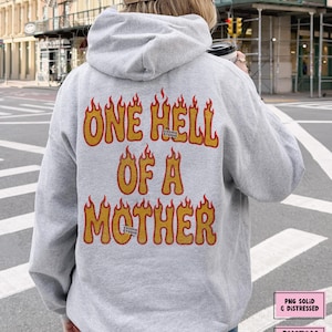 May include: A light gray hoodie with the text "ONE HELL OF A MOTHER" in orange and yellow flames. The hoodie has a drawstring hood and is made of a soft material. The Cosmic Studio logo is also visible.