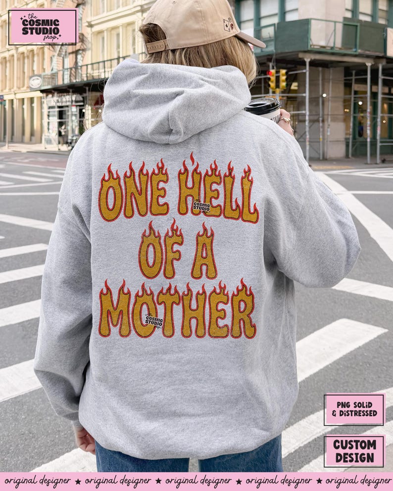 May include: A light gray hoodie with the text "ONE HELL OF A MOTHER" in orange and yellow flames. The hoodie has a drawstring hood and is made of a soft material. The Cosmic Studio logo is also visible.