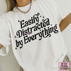May include: A white t-shirt with the black text "Easily Distracted by Everything". The design includes star accents. The Cosmic Studio shop logo is visible. The shirt has a crew neck and short sleeves. A digital download is available.