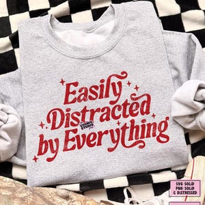 May include: A grey sweatshirt featuring the red text "Easily Distracted by Everything". The design includes star accents and the Cosmic Studio logo. The sweatshirt has a crew neck and long sleeves.