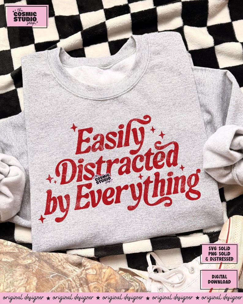 May include: A grey sweatshirt featuring the red text "Easily Distracted by Everything". The design includes star accents and the Cosmic Studio logo. The sweatshirt has a crew neck and long sleeves.