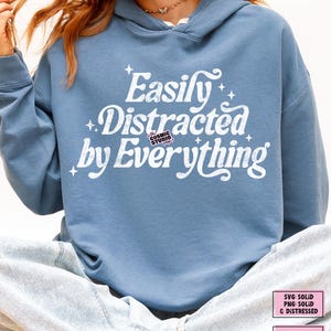 May include: A pale blue hoodie featuring the white text "Easily Distracted by Everything". The hoodie has a drawstring hood and is made of a soft material. The Cosmic Studio shop logo is also visible.