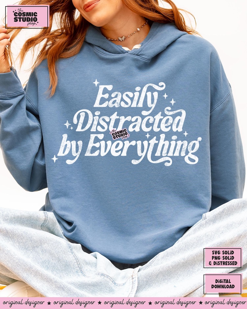 May include: A pale blue hoodie featuring the white text "Easily Distracted by Everything". The hoodie has a drawstring hood and is made of a soft material. The Cosmic Studio shop logo is also visible.