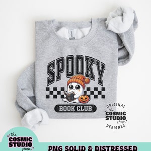 May include: A grey sweatshirt with a distressed graphic design featuring a ghost wearing a beanie and reading a book. The text "Spooky Book Club" is below the ghost. The text "Original the Cosmic Studio shop. Designer" is on the right side of the graphic.