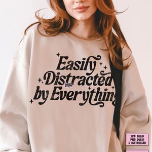 May include: A beige sweatshirt featuring the phrase "Easily Distracted by Everything" in black, stylized lettering. The design incorporates star motifs. Additional text indicates the design is available as SVG and PNG files, and as a digital download.