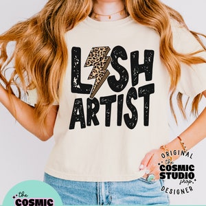Puede incluir: Un dise&ntilde;o gr&aacute;fico en blanco y negro desgastado con el texto "LASH ARTIST" y un rayo de impresi&oacute;n de leopardo. El dise&ntilde;o est&aacute; en una camiseta beige. El texto "ORIGINAL the COSMIC STUDIO shop DESIGNER" est&aacute; en un c&iacute;rculo blanco con un contorno negro. El texto "PNG DISTRESSED" est&aacute; en un rect&aacute;ngulo rosa con un contorno blanco.