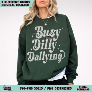 May include: A dark green sweatshirt with the text "Busy Dilly Dallying" in a white, vintage-style font. The sweatshirt has a crew neck and long sleeves. The design includes the text "the COSMIC STUDIO shop."