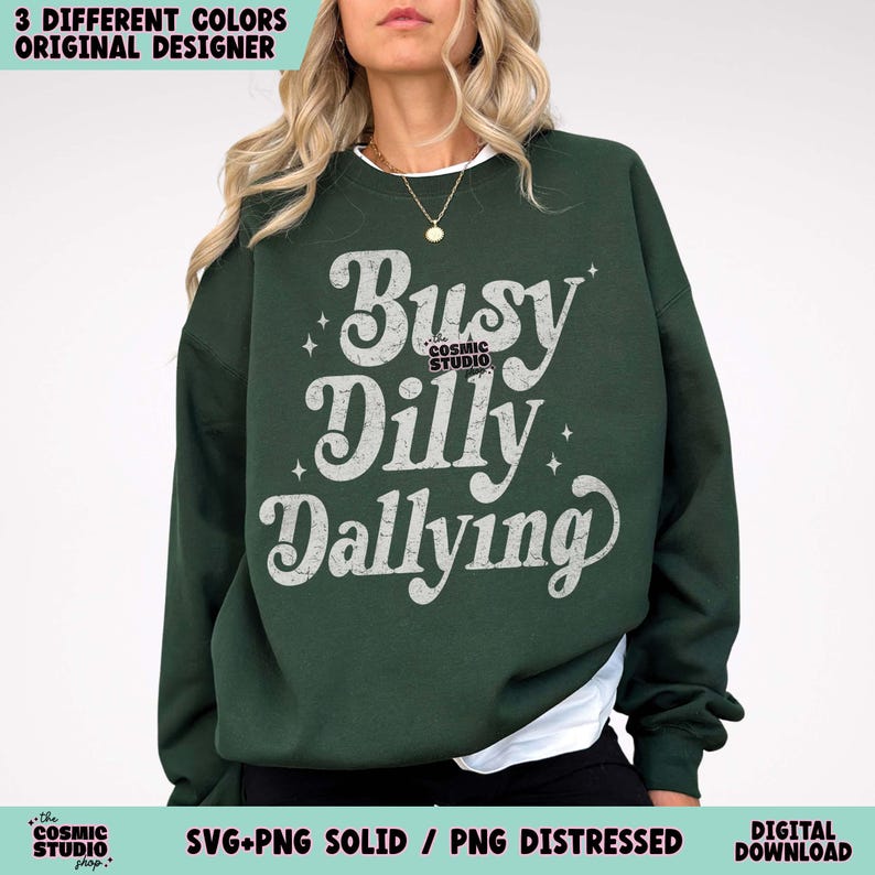 May include: A dark green sweatshirt with the text "Busy Dilly Dallying" in a white, vintage-style font. The sweatshirt has a crew neck and long sleeves. The design includes the text "the COSMIC STUDIO shop."