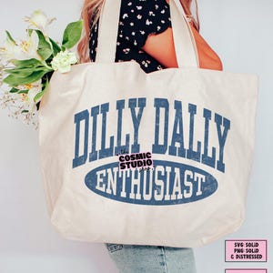 K&ouml;nnte beinhalten: Eine gro&szlig;e cremewei&szlig;e Tragetasche aus Segeltuch mit dem blauen Schriftzug DILLY DALLY ENTHUSIAST in einer abgenutzten College-Schriftart. Das Design enth&auml;lt eine blaue ovale Form hinter dem Wort ENTHUSIAST und den Text the COSMIC STUDIO shop.