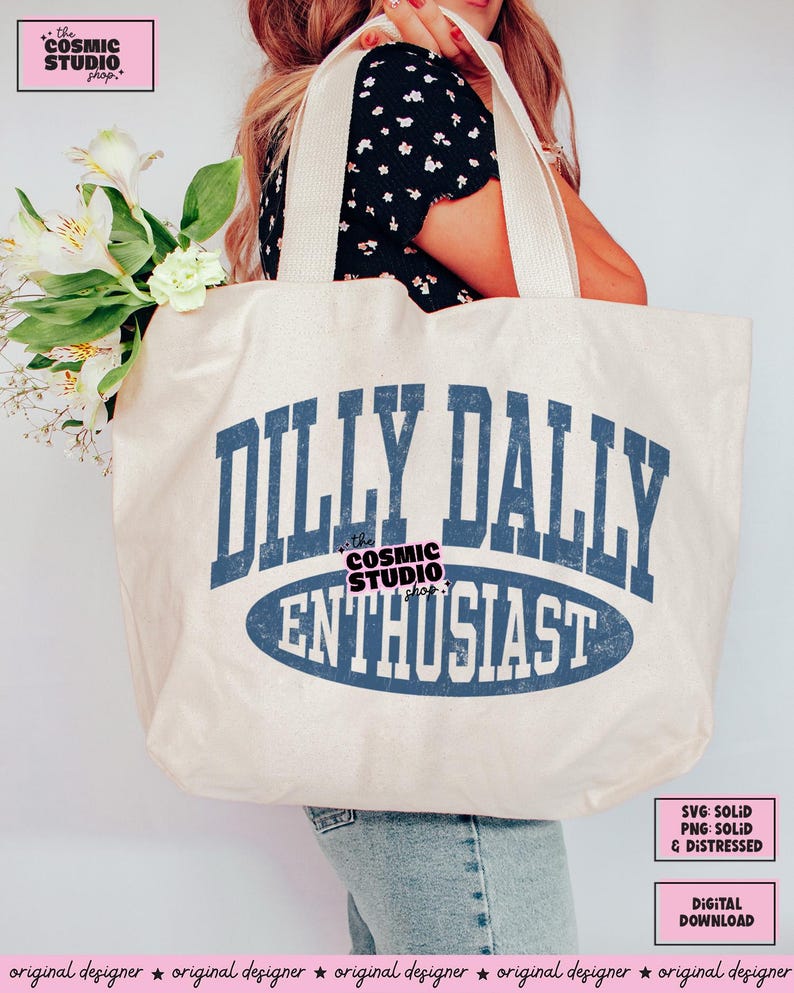 K&ouml;nnte beinhalten: Eine gro&szlig;e cremewei&szlig;e Tragetasche aus Segeltuch mit dem blauen Schriftzug DILLY DALLY ENTHUSIAST in einer abgenutzten College-Schriftart. Das Design enth&auml;lt eine blaue ovale Form hinter dem Wort ENTHUSIAST und den Text the COSMIC STUDIO shop.