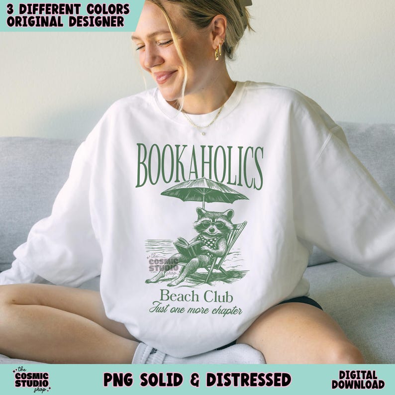 May include: White sweatshirt with a green raccoon reading a book under an umbrella. The text "Bookaholics Beach Club Just one more chapter" is printed on the shirt.