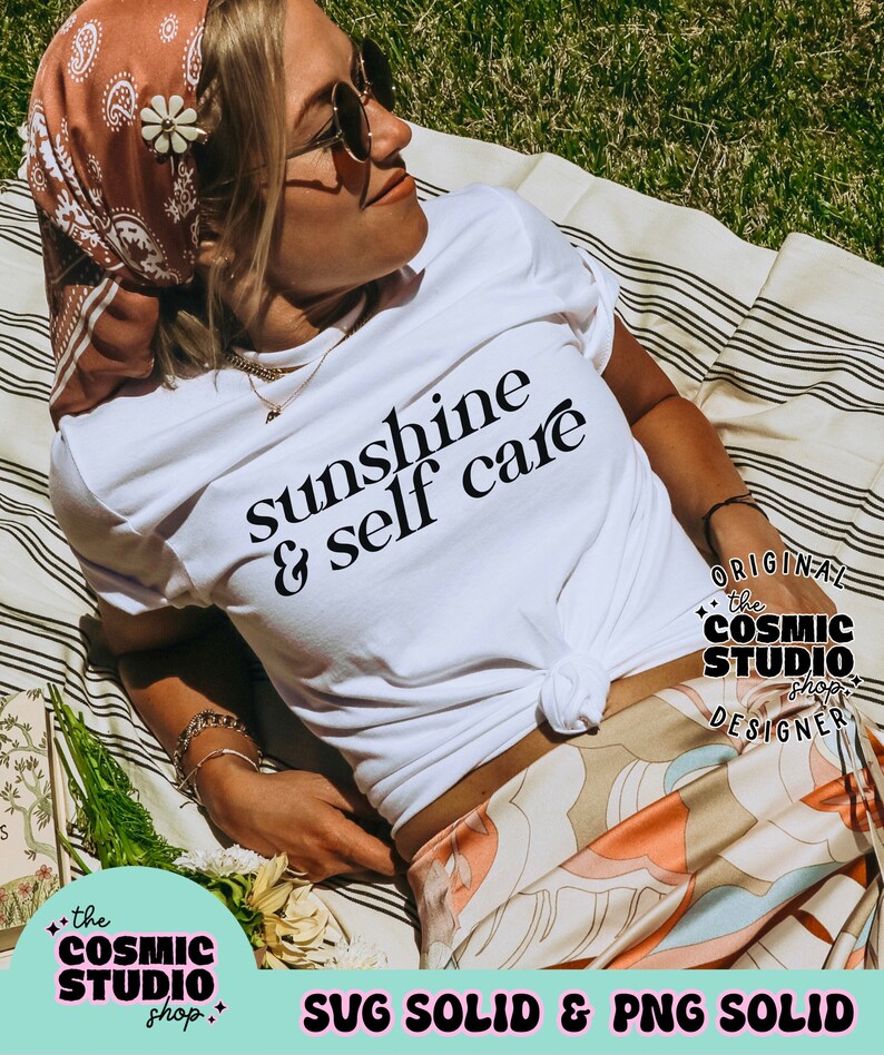 May include: A white t-shirt with black text that reads "sunshine & self care". The shirt is worn by a person lying on a striped blanket. The image includes a logo for "the cosmic studio shop" in the bottom left corner and a logo for "the cosmic studio shop designer" in the bottom right corner.