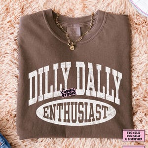 K&ouml;nnte beinhalten: Ein braunes T-Shirt mit Rundhalsausschnitt und wei&szlig;em, verwaschenem Schriftzug im College-Stil, der DILLY DALLY ENTHUSIAST lautet. Das T-Shirt ist mit einer goldfarbenen Kette mit einem kleinen herzf&ouml;rmigen Anh&auml;nger mit dem Buchstaben M kombiniert.