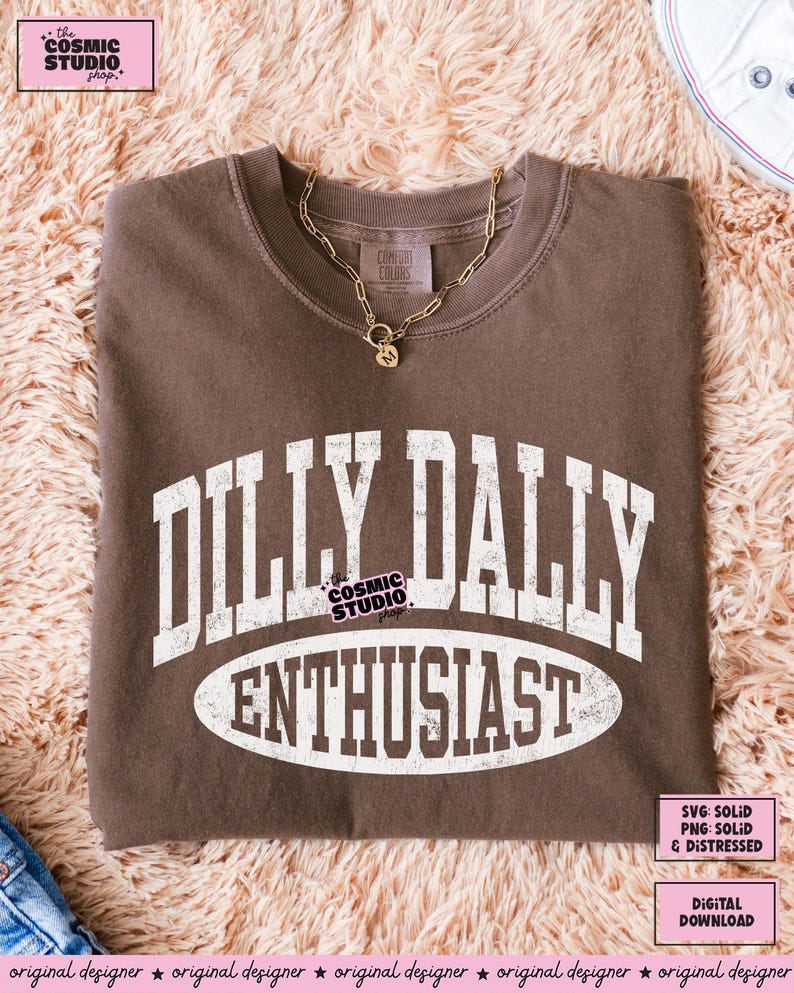 K&ouml;nnte beinhalten: Ein braunes T-Shirt mit Rundhalsausschnitt und wei&szlig;em, verwaschenem Schriftzug im College-Stil, der DILLY DALLY ENTHUSIAST lautet. Das T-Shirt ist mit einer goldfarbenen Kette mit einem kleinen herzf&ouml;rmigen Anh&auml;nger mit dem Buchstaben M kombiniert.