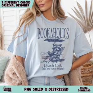 May include: A light blue t-shirt featuring a graphic of a raccoon reading a book on a beach chair under an umbrella. The text "BOOKAHOLICS" is above the graphic and "Beach Club Just one more chapter" is below the graphic.