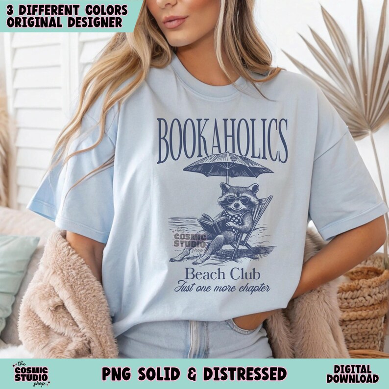 May include: A light blue t-shirt featuring a graphic of a raccoon reading a book on a beach chair under an umbrella. The text "BOOKAHOLICS" is above the graphic and "Beach Club Just one more chapter" is below the graphic.
