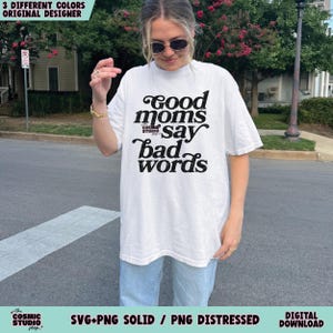 May include: White t-shirt with the text "Good moms say bad words" in a black, stylized font. The shirt is part of the "Cosmic Studio" shop collection. The image also includes text indicating the availability of SVG and PNG files.