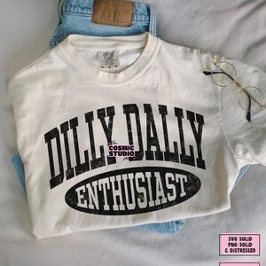K&ouml;nnte beinhalten: Ein cremefarbenes Kurzarm-T-Shirt mit dem schwarzen Schriftzug DILLY DALLY ENTHUSIAST in einer abgenutzten College-Schriftart. Das Wort ENTHUSIAST ist in einem schwarzen Oval eingeschlossen. Das T-Shirt hat ein Comfort Colors Etikett und blaue Jeans.