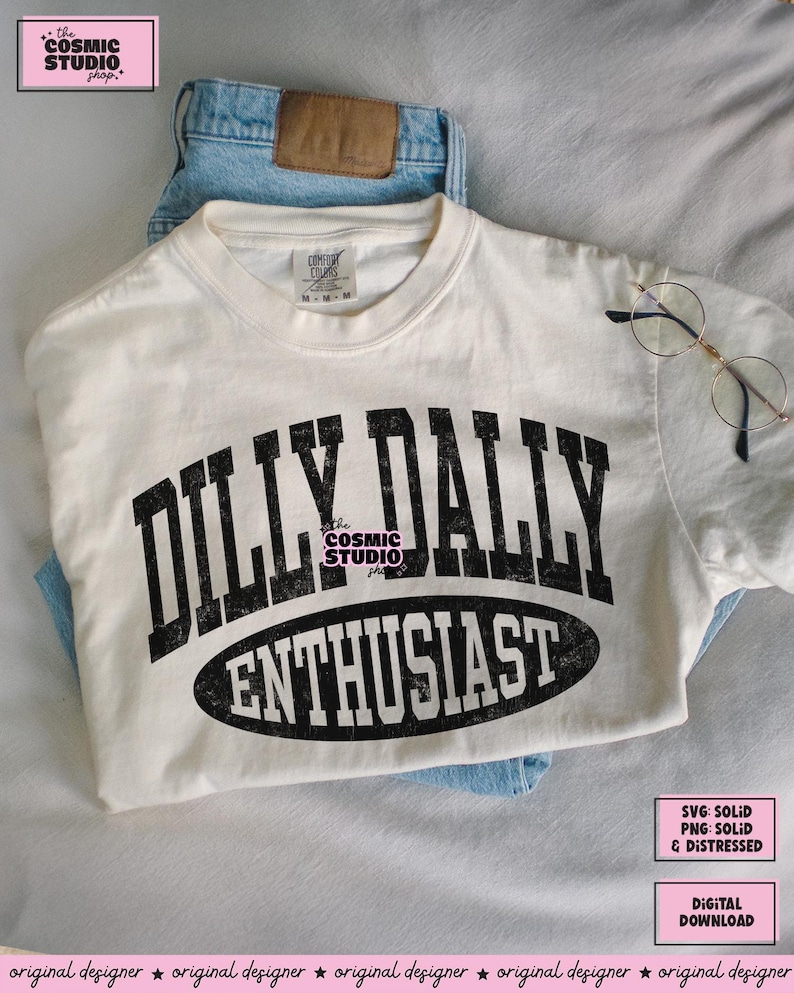 K&ouml;nnte beinhalten: Ein cremefarbenes Kurzarm-T-Shirt mit dem schwarzen Schriftzug DILLY DALLY ENTHUSIAST in einer abgenutzten College-Schriftart. Das Wort ENTHUSIAST ist in einem schwarzen Oval eingeschlossen. Das T-Shirt hat ein Comfort Colors Etikett und blaue Jeans.