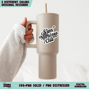 May include: A beige tumbler with a black and white sticker that says "Slow Mornings Club". The tumbler has a straw and a handle.